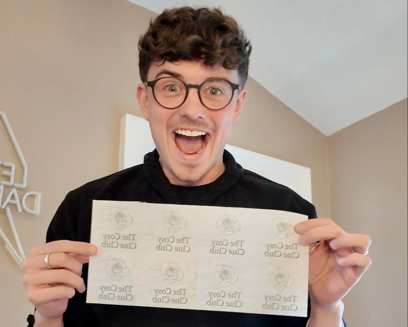 A man with curly brown hair and glasses, wearing a black t-shirt, holds up a sheet of white stickers printed with the text 'THE ONLY' in a circular design. The background shows a beige wall and a white ceiling.